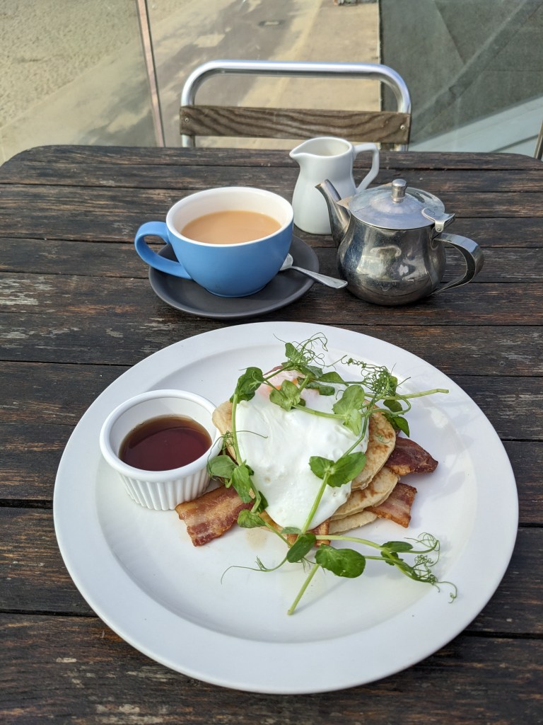 Photo of American-style pancakes with egg and bacon and maple syrup on the side on a white china plate. The plate is on a wooden table and above the plate is a cup of rooibos tea, a teapot and a milk jug. Chineside cafe in Bournemouth.
