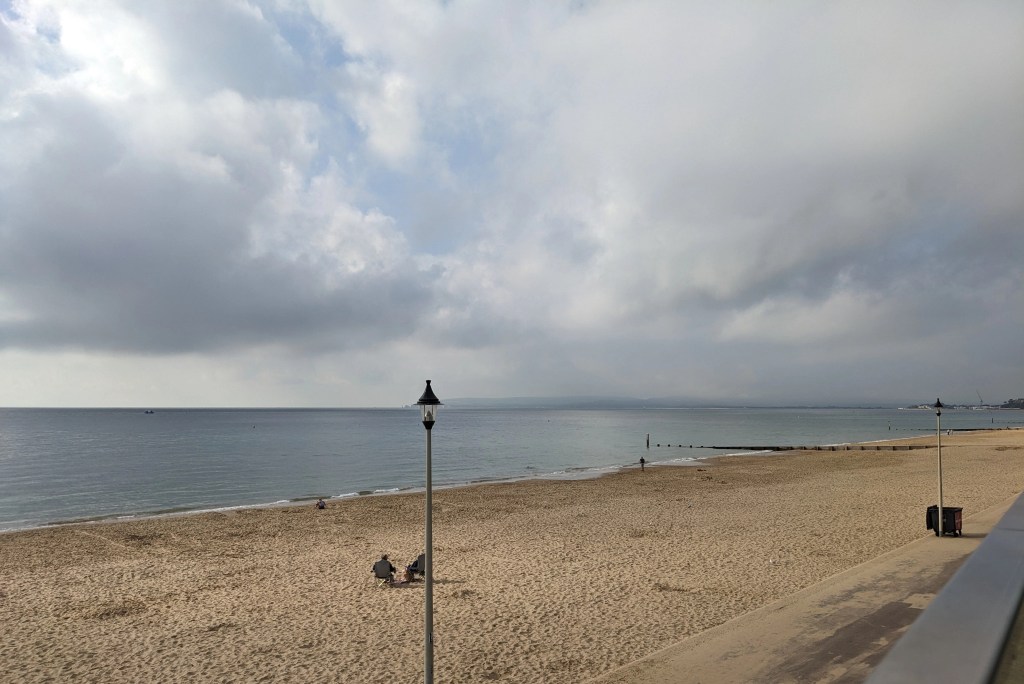 Photo of the seaside in Bournemouth. White clouds in the sky, blue calm sea, yellow sandy beach with two small figures sat on chairs. 