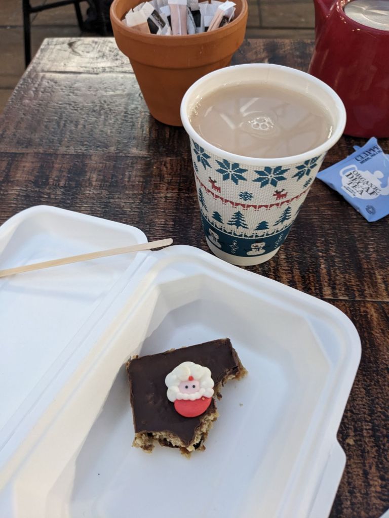 A photo of a part eaten flapjack in a white box. The flapjack is topped with chocolate and has a Santa face decoration on. A takeaway cup of weak tea is next to it. The cup is has a printed Christmas design on. All on a brown wooden table top.